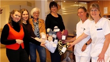 Sechs Frauen stehen in einem Krankenhausraum und halten bunte herzförmige Kissen in den Händen. Zwei der Frauen tragen Pflegekleidung, die anderen Alltagskleidung. Es werden Kissen übergeben.