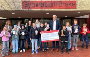 Eine Gruppe von Schülerinnen, Schülern und Erwachsenen steht vor dem Eingang des Gymnasiums Brake. Sie halten Edelstahltrinkflaschen und Geschenkboxen in den Händen. Ein Mann in einem Anzug präsentiert gemeinsam mit den Kindern einen großen Spendenscheck über 1.500 Euro. Der Hintergrund zeigt den Eingang des Schulgebäudes mit dem Schild "Gymnasium Brake".