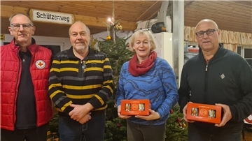 Vier Personen stehen nebeneinander vor einem geschmückten Weihnachtsbaum in einem Raum mit Holzdecke. Links trägt eine Person eine rote Jacke mit dem Logo des Deutschen Roten Kreuzes.