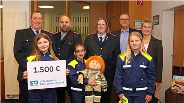  Drei Kinder in blauen und gelben Feuerwehruniformen stehen in der vorderen Reihe, eines hält ein Plakat mit der Aufschrift '1.500 € Volksbank im Elbe-Weser-Dreieck', ein anderes eine Stoffpuppe in Feuerwehrkleidung. Dahinter stehen fünf Erwachsene, darunter Männer in Feuerwehruniformen und zwei Personen in Zivilkleidung.