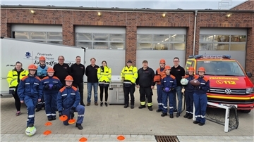 Gruppenfoto vor dem Feuerwehrhaus Wulsdorf: Mitglieder der Jugendfeuerwehr in blauer Schutzkleidung und orangenen Helmen, sowie Betreuer und Unterstützer in schwarzer oder gelber Kleidung stehen gemeinsam vor einem Feuerwehrfahrzeug und einem Anhänger. Im Vordergrund liegen Sportgeräte wie Bälle, Hütchen und ein kleines Tor – im Zentrum ein neuer Edelstahlgrill