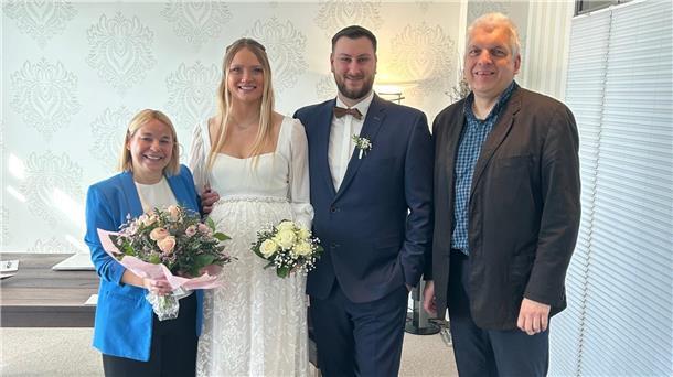 Vier Personen posieren fröhlich für ein Foto bei einer standesamtlichen Hochzeit. In der Mitte stehen das Brautpaar – die Braut in weißem Kleid mit Blumenstrauß, der Bräutigam im blauen Anzug mit Fliege. Links steht eine Frau mit blauem Blazer und einem weiteren Blumenstrauß, rechts ein Mann im dunklen Sakko.