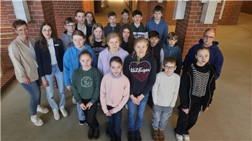 Eine Gruppe von rund 20 Personen, bestehend aus Schülerinnen und Schülern sowie zwei Frauen, steht im Flur eines Verwaltungsgebäudes vor einer Backsteinwand.