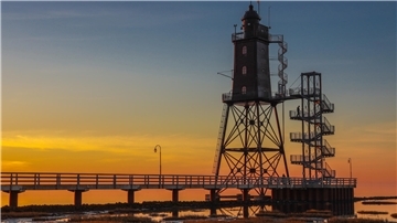**ALT-Text:**  
Historischer Leuchtturm Obereversand in Dorum-Neufeld bei Sonnenuntergang, mit Steg und Treppe im Vordergrund und farbenfrohem Himmel im Hintergrund.