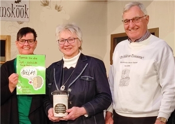 Drei lächelnde Personen stehen nebeneinander. Die Frau links hält ein grünes Schild mit der Aufschrift „Spende für die Igel-Nothilfe“ und dem Betrag 1079,81 Euro. Die Frau in der Mitte hält eine Spendenbox. Der Mann rechts trägt einen weißen Pullover mit dem Aufdruck "De Lüüd von Ossenturm".