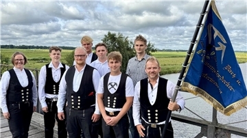 Gruppe von acht Personen in traditioneller Zunftkleidung und moderner Kleidung steht auf einer Brücke vor einer ländlichen Flusslandschaft. Eine Person hält eine blau-goldene Zunftfahne des Dachdeckerhandwerks.