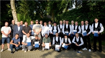 Gruppenfoto von jungen Männern, teils in traditioneller Handwerkskleidung, teils in Freizeit- oder festlicher Kleidung, viele mit Zeugnismappen in der Hand. Die Gruppe steht im Freien vor grünem, bewaldetem Hintergrund