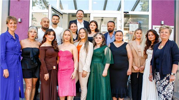 Gruppenfoto von 16 Personen in festlicher Kleidung vor einem Gebäude mit Glasdach und lilafarbener Wand. Männer und Frauen posieren gemeinsam für ein Bild.