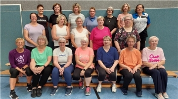 Gruppenfoto der Frauen-Fitnessgruppe des TSV Hollen mit 21 Teilnehmerinnen in Sportkleidung, aufgenommen in einer Turnhalle anlässlich des 50-jährigen Jubiläums. Die Frauen stehen und sitzen auf Bänken vor einer blauen Wand.