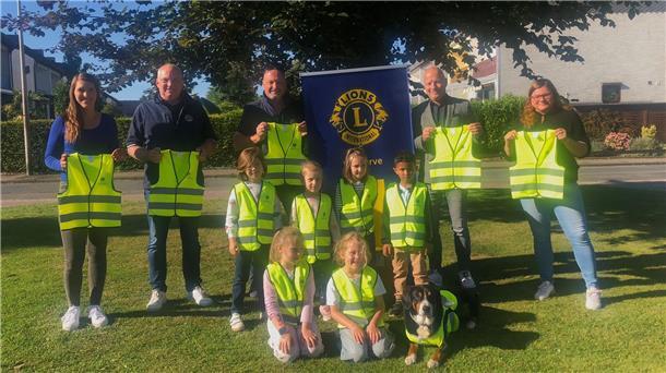 Mehrere Erwachsene und Kinder stehen und knien auf einer Wiese, alle tragen oder halten leuchtend gelbe Sicherheitswesten. Hinter ihnen ist ein Banner des Lions Clubs zu sehen. Auch ein Hund trägt eine Warnweste.