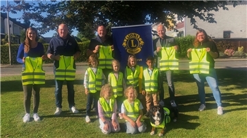 Mehrere Erwachsene und Kinder stehen und knien auf einer Wiese, alle tragen oder halten leuchtend gelbe Sicherheitswesten. Hinter ihnen ist ein Banner des Lions Clubs zu sehen. Auch ein Hund trägt eine Warnweste.