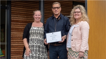 Drei Personen stehen lächelnd vor einem modernen Holzgebäude. Die mittlere Person hält eine gerahmte Spendenurkunde über 1.500 Euro der Proske-Stiftung in den Händen.