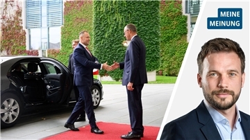 Zwei Männer geben sich zur Begrüßung auf einem roten Teppich die Hand. Ein schwarzer Wagen steht hinter einem der Männer. 