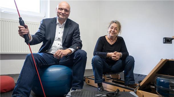 Ein Mann im Anzug sitzt auf einem blauen Gymnastikball und hält ein Trainingsgerät mit rotem Widerstandsband. Neben ihm sitzt eine Frau in schwarzem Oberteil und Jeans auf einem Sportgerät.