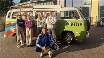 Ein Mann hockt vor einem Bulli des Radio Senders Bremen 1. Hinter ihm stehen vier lächelnde junge Leute. 