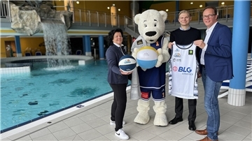 Drei Personen und ein Maskottchen in Eisbärenkostüm stehen am Beckenrand der Moor-Therme. Sie halten Basketbälle und ein Trikot, im Hintergrund ist ein Wasserfall zu sehen.