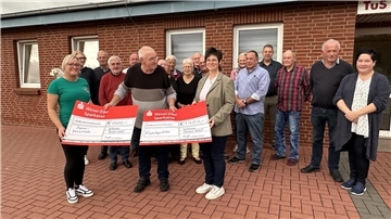 Gruppenfoto von einer Spendenübergabe. Im Vordergrund halten drei Personen zwei symbolische Spendenschecks in die Höhe.