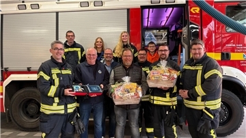 Defibrillator für die Ortsfeuerwehr Langen Gruppenbild vor einem Einsatzfahrzeug der Feuerwehr Langen. Menschen halten einen Defibrillator und Präsentkörbe in den Händen.