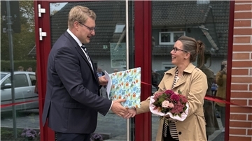 Schiffdorf weiht neue Kindertagesstätte IV ein Ein Mann im Anzug überreicht einer Frau mit Blumenstrauß vor einem Gebäude ein Geschenk. Beide lächeln, im Hintergrund ist ein rotes Band zur Eröffnung zu sehen.