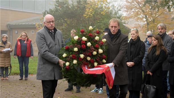 Zwei Männer tragen einen Kranz mit roten und weißen Blüten. Im Hintergrund stehen Menschen.