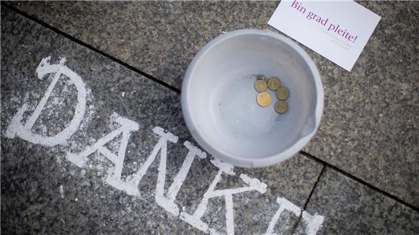 Ein Sammelbehälter mit wenigen Münzen steht auf dem Boden neben einer Postkarte mit der Aufschrift "Bin grad pleite!" und dem Kreide-Schriftzug "Danke"
