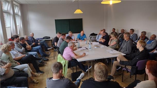 In der Bildmitte sitzen elf Menschen um einen Tisch herum, und an den Wänden sitzen Besucher auf langen Stuhlreihen.