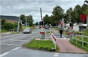 Ein Straße, auf der ein Auto fährt. Bahnschranken sind zu sehen, ebenso wie ein Fußgänger.