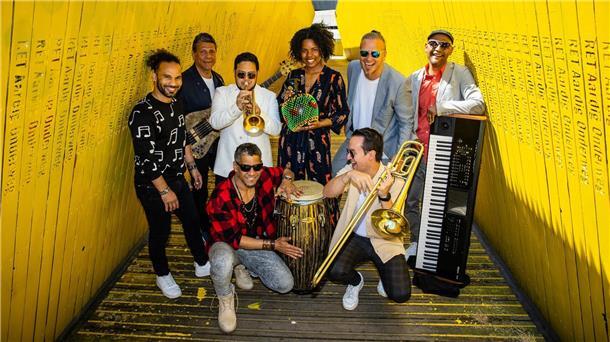 Die achtköpfige Band „CaboCubaJazz“ posiert fröhlich in einem gelben Durchgang. Die Musiker halten Instrumente wie Trompete, Conga, Posaune und Keyboard.
