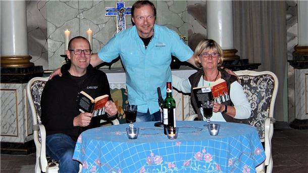 Eine Frau und ein Mann sitzen an einem Tisch. Sie halten jeweils ein Buch in der Hand. Ein weiterer Mann steht zwischen den beiden.
