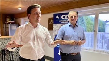 Ein Mann mit weißem Hemd gestikuliert mit beiden Händen, daneben steht ein Mann mit blauem Hemd, der die Hände vor dem Bauch verschränkt. 