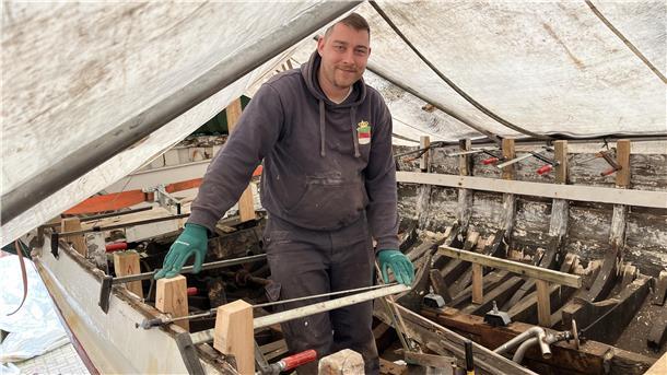 Mann steht auf einem Boot unter einer Plane. Christian Schwan macht ein Börteboot wieder fit. Zur Sail im August soll es spätestens fahren.