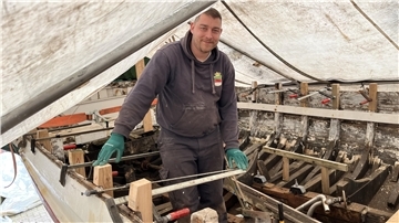 Mann steht auf einem Boot unter einer Plane. Christian Schwan macht ein Börteboot wieder fit. Zur Sail im August soll es spätestens fahren.