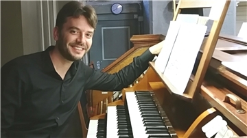 Ein lächelnder Mann in dunklem Hemd sitzt an einer historischen Orgel und blickt in die Kamera. Seine rechte Hand ruht auf einem Notenständer mit geöffnetem Notenbuch.