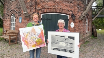 Zwei Frauen stehen vor einer Mühle. Sie halten jeweils ein großes Bild in den Händen.