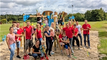 Gruppe von Erwachsenen und Kindern vor einem neu aufgebauten Spielplatz mit Klettergerüst und Rutsche auf einem Sandplatz, im Hintergrund ein Sportplatz und Bäume. Einige tragen rote Vereinsshirts, mehrere Kinder spielen oder werden getragen.