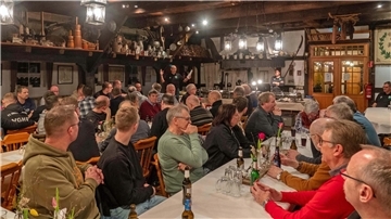 Ein geselliges Treffen in einer rustikalen Gaststube mit vielen Personen, die an langen Tischen sitzen. Eine Person steht vorne und hält eine Rede, während die Gäste aufmerksam zuhören. Die Wände und Decken sind mit alten Haushaltsgegenständen dekoriert, und die Beleuchtung besteht aus traditionellen Laternen. Auf den Tischen stehen Getränke und Blumendekorationen.