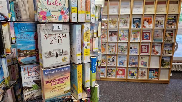 Für gemütliche Lesestunden: Buchtipps aus der Zevener Bibliothek Ein Bücherständer im Vordergrund und ein Regal mit Magazinen im Hintergrund.