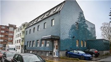 Mehrfamilienhaus mit blau-grauer Fassade im Regen, davor und daneben geparkte Autos.