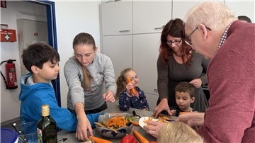 Mehrere Kinder, Erwachsene und ein älterer Mann schälen gemeinsam Karotten an einem Tisch in einer Küche. Sie wirken konzentriert und fröhlich beim gemeinsamen Kochen.