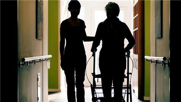 Eine Pflegekraft (l.) begleitet die Bewohnerin eines Altenheims mit Rollator beim Gang über den Flur.