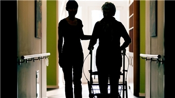 Eine Pflegekraft (l.) begleitet die Bewohnerin eines Altenheims mit Rollator beim Gang über den Flur.