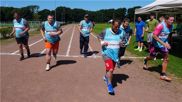 **ALT-Text:**
Mehrere Männer starten bei sonnigem Wetter auf einer Tartanbahn zu einem Lauf im Rahmen des Deutschen Sportabzeichens. Sie tragen blaue Startwesten mit Nummern. Im Hintergrund stehen Helfer und Zuschauer an einem weißen Pavillon.