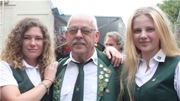 Drei Mitglieder des Schützenvereins Bramstedt beim Jubiläumsschützenfest: Ein älterer Mann in Uniform mit Orden steht zwischen zwei jungen Frauen in grüner Vereinskleidung, alle lächeln in die Kamera.