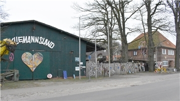 Eine grüne Scheune auf der linken Seite und Bäume und ein roter Klinkerbau auf der rechten Seite.