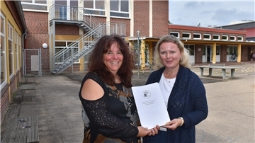 Schulleiterin Gerda Vogel und Lehrervertreterin Daniele van de Velde stehen auf dem Schulhof der Grundschule Butjadingen und halten gemeinsam ein Konzeptpapier in Händen
