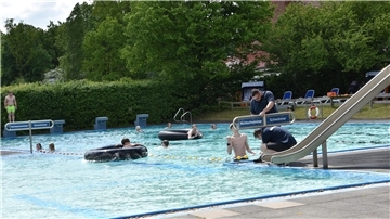 Ein Freibad mit mehreren Personen im Wasser. Einige Menschen benutzen große Schwimmringe, andere schwimmen oder stehen im flachen Wasser. Rechts im Bild befindet sich eine Wasserrutsche. Am unteren Ende montieren zwei Männer ein Schild, das das Becken in Nichtschwimmer und Schwimmer aufteilt. 