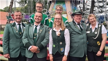 Eine Gruppe von acht Personen, bestehend aus Männern und Frauen, posiert lächelnd für ein Gruppenfoto vor einem Karussell. Die vordere Reihe trägt traditionelle grüne Uniformen und Westen mit Abzeichen und Schützenorden, während zwei junge Männer in der hinteren Reihe grün-weiße Sportshirts tragen.
