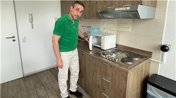 Ein Mann mit Brille, grünem Polo-Hemd und weißer Hose steht in einer Küche. Er öffnet die Mikrowelle, die links neben dem Herd steht. Darüber die Abzugshaube. Die Küche besteht aus hellbraunen Holzschränken.