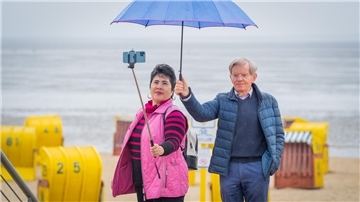 Eine Frau und ein Mann stehen am Strand von Duhnen. Sie hält ein Smartphone auf einem Selfiestick, er den Regenschirm.