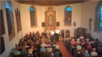 Zwei Männer stehen vor dem Kirchenaltar. Die Bänke in der Kirche sind voll besetzt. 